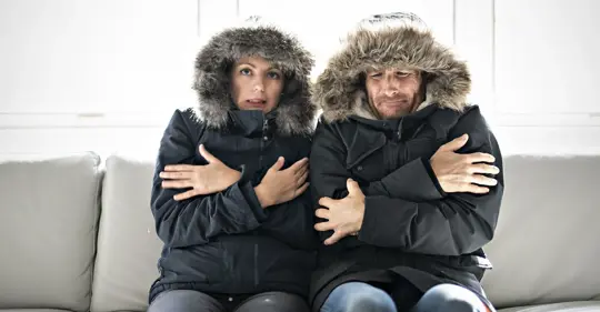 A man and woman in winter coats sit on a couch shivering